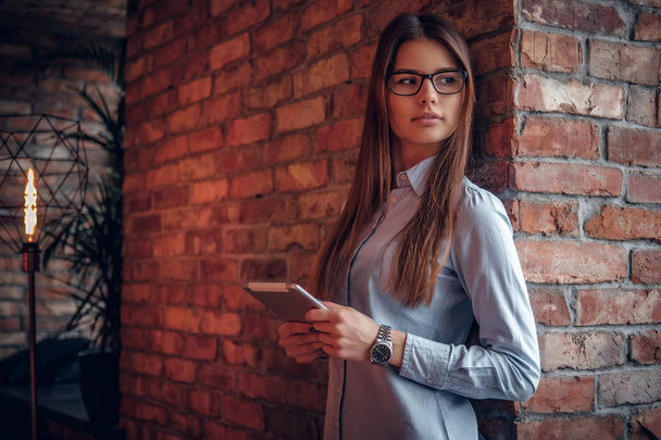Smart girl vêtue d'une chemise élégante tenant la tablette tout en s'appuyant sur un mur de briques dans la chambre avec un intérieur loft
  - Photo, image