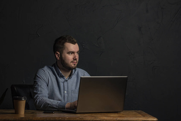 Jovem empresário trabalhando em seu laptop na mesa de madeira. Espaço para SMS. Fundo escuro. Espaço para texto
 - Foto, Imagem