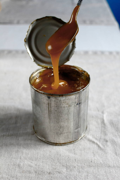 Leche condensada hervida tradicional casera en una lata vertida de una cuchara sobre la mesa
 - Foto, imagen