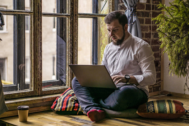 Giovane uomo che lavora con il computer portatile a casa seduto sul pavimento contro la finestra
 - Foto, immagini