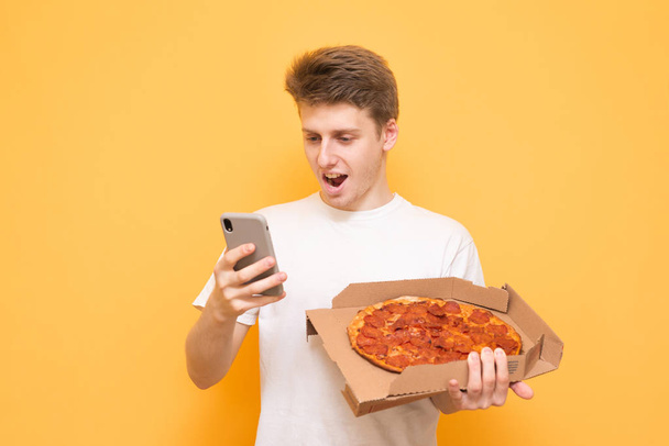 Happy young man in a white T-shirt holds a box of pizza and uses a smartphone on a yellow background. Young man with a smartphone and pizza in his hands is isolated on a yellow background. - Photo, image