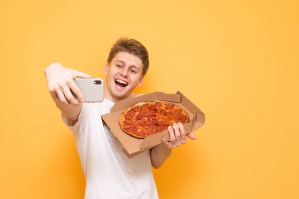 Happy guy takes selfie with a box of pizza on a yellow background. Young man takes a photo of a pizza on a smartphone, isolated on a yellow background. Copyspace - Photo, Image