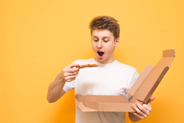 Hungry guy with pizza in his hands is isolated on a yellow background. Emotional young man in a white T-shirt holds in a box and a piece of pizza, looks at an appetizing pizza with a hungry look. - Photo, Image