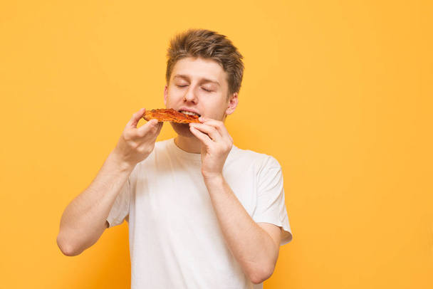 Young man eats a piece of appetizing pizza on a yellow background. Hungry guy holds a piece of pizza in his hands, looks at him and is going to eat it, isolated on a yellow - Valokuva, kuva