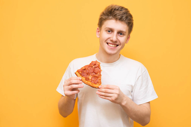 Portrait of a smiling guy dressed in a white T-shirt isolated on a yellow background, holding a piece of pizza in his hands and looking into the camera. Teenager and fast food. Copyspace - Valokuva, kuva
