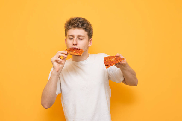 Guy dressed in a white T-shirt holds in his hands a piece of pizza and eats fast food on a yellow background. Young man eats a piece of pizza with his eyes closed, isolated on a yellow background. - Photo, Image