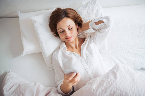 A top view of young woman lying in bed indoors in a bedroom, using smartphone. - Foto, Imagen