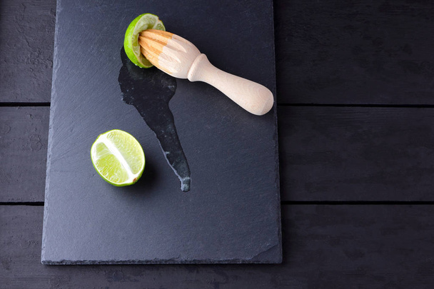 Lime halves and wooden juicer on slate board. Lime on black background. Fresh lime juice flows over the stone board. Healthy food - 写真・画像