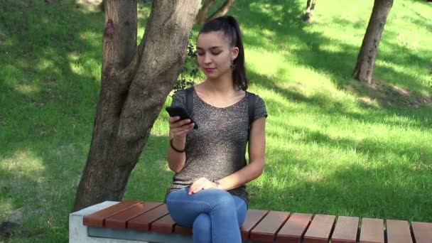 Young beautiful girl is sitting on a bench in the park and browsing the social networking tape in her phone. - Filmmaterial, Video
