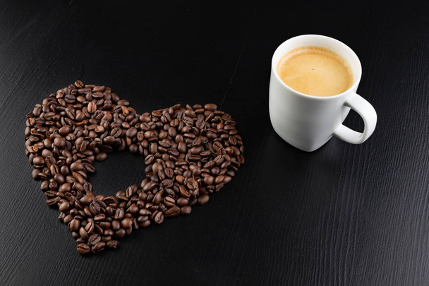 Granos de café tostados dispuestos en forma de corazón sobre la mesa. Sabroso café fragante en una taza blanca. Fondo oscuro
. - Foto, Imagen