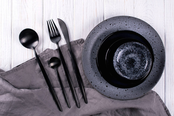Ensemble de couverts en porcelaine et assiettes grises et noires, serviette en lin sur une table en bois blanc. Pose plate
 - Photo, image