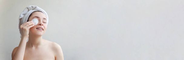 Schönheitsporträt einer lächelnden Frau in Handtuch auf dem Kopf mit weicher, gesunder Haut, die Make-up entfernt, mit Wattepad isoliert über weißem Hintergrund. Reinigungs-Wellness-Entspannungskonzept. Banner - Foto, Bild