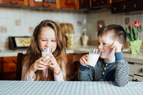 Children drink milk and have fun in the kitchen at the morning. Sister and brother sitting at the table at home - Фото, изображение