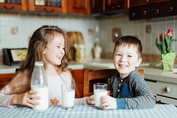Kinder trinken Milch und amüsieren sich morgens in der Küche. Schwester und Bruder sitzen zu Hause am Tisch und Schwester gießt Milch in ein Glas - Foto, Bild
