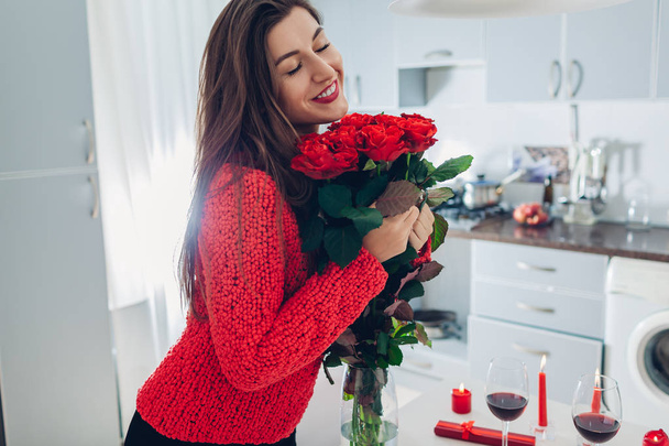 Young woman found bouquet of roses with candle, wine and gift box on kitchen. Happy girl holding flowers. Valentines day surprise - Foto, Imagem