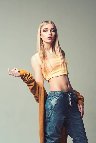 Blond girl with straight hair isolated on gray background. Young model in jeans posing in studio - Фото, изображение