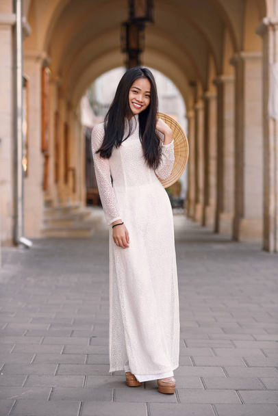 Beautiful happy asian girl dressed in Ao Dai dress with vietnamese conical hat against arches background. - Zdjęcie, obraz