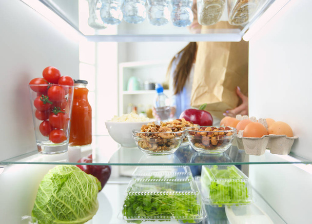 Portrait of female standing near open fridge full of healthy food, vegetables and fruits. - Photo, image