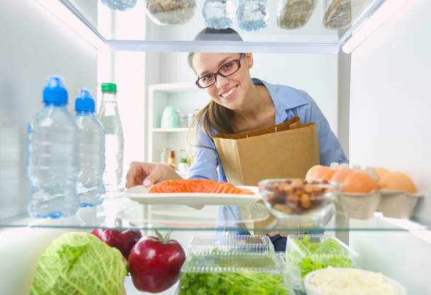 Portrait of female standing near open fridge full of healthy food, vegetables and fruits. - Photo, Image