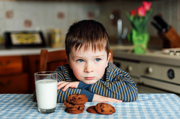 Ένα μικρό αγόρι ποτά γάλακτος και τρώει μπισκότα στην κουζίνα το πρωί. - Φωτογραφία, εικόνα