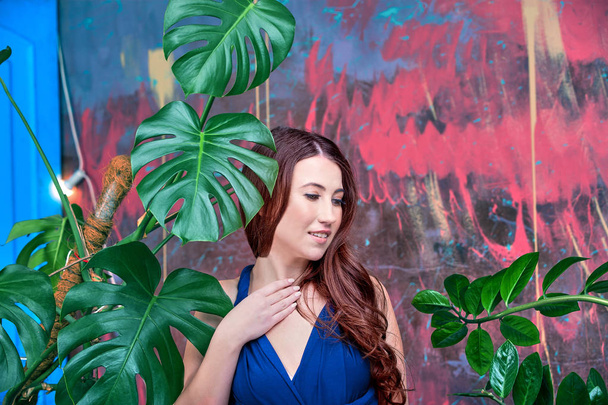 Close up portrait of a young brunette lady, wearing blue dress. The girl posing, looking down. Green palm leaves in the background. - Fotografie, Obrázek