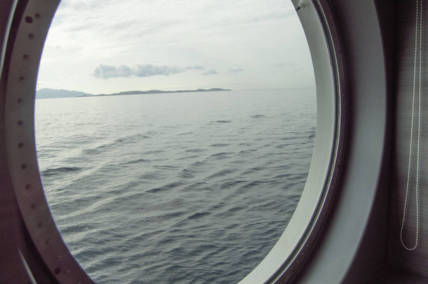 Круглый иллюминатор на круизном лайнере, вид изнутри через окно на побережье и море, восход солнца на море, крупный план
. - Фото, изображение
