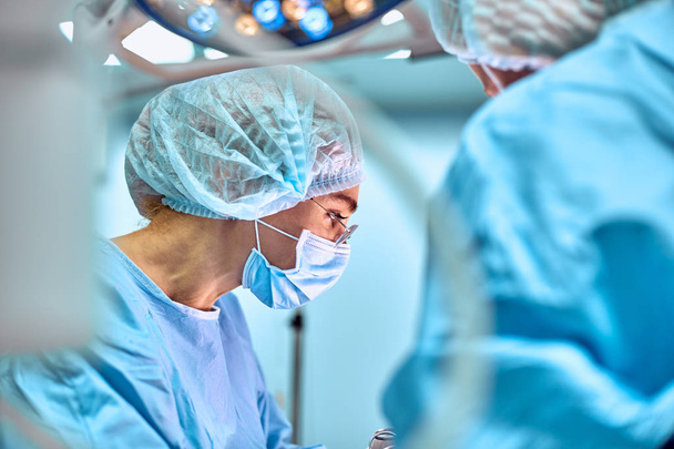 Retrato de perto de uma jovem médica cirurgiã usando máscara protetora e chapéu durante a operação. Saúde, educação médica, conceito de cirurgia
 - Foto, Imagem