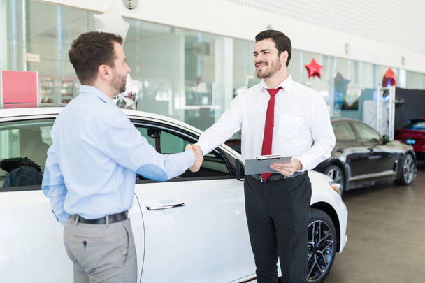 Hymyilevä myynti asiantuntija toivottaa puolivälissä aikuinen asiakas auton showroom
 - Valokuva, kuva