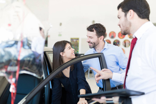 Inhoud van de mannelijke en vrouwelijke klanten kijken elkaar terwijl het controleren van auto interieurs op showroom - Foto, afbeelding