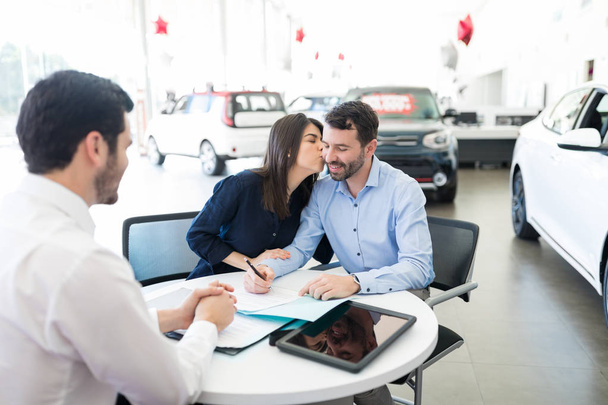 Iloinen nainen suutelee miestä allekirjoittamalla osto- ja myyntisopimuksen autokaupassa
 - Valokuva, kuva
