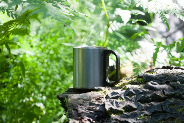 A marching metal mug with a hot drink in a wild forest on a snag. Summer sunny day - Фото, изображение