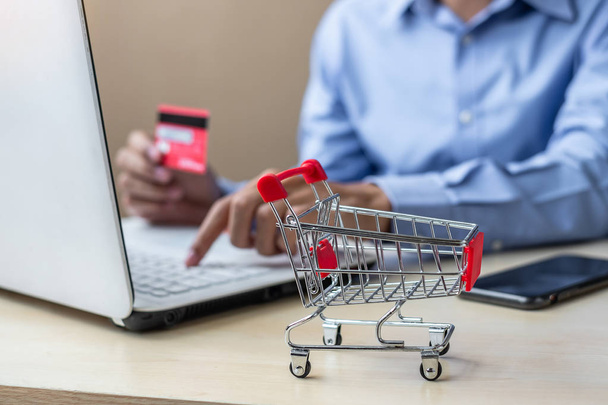 Nahaufnahme Mini-Warenkorb, asiatische Geschäftsmann mit Kreditkarte und Laptop für den Online-Einkauf während der Bestellung. Internet, Technologie, E-Commerce und Online-Zahlungskonzept - Foto, Bild