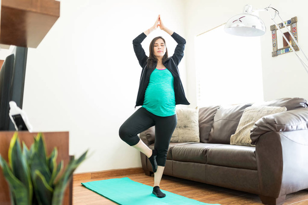 Mid adult pregnant woman performing tree pose on yoga mat at home - Foto, imagen