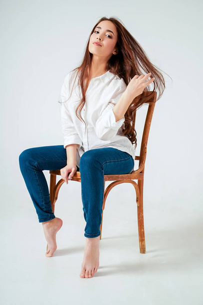 Beauty fashion portrait of smiling sensual asian young woman with dark long hair in white shirt sitting on chair on white background - Valokuva, kuva