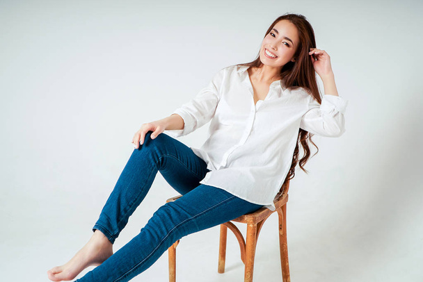 Красивий портрет моди усміхненої чуттєвої азіатської молодої жінки з темним довгим волоссям у білій сорочці, що сидить на стільці на білому тлі
 - Фото, зображення