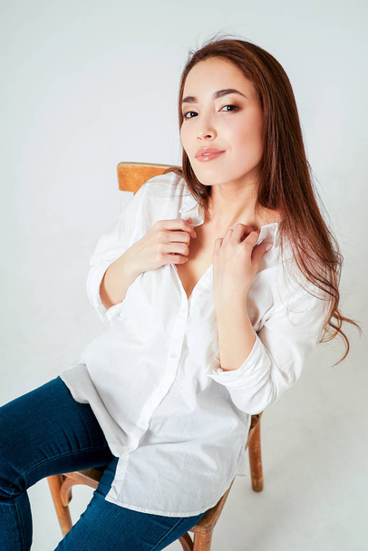Mosolygó érzéki ázsiai fiatal nő ül a széken a fehér háttér előtt fehér ing-sötét hosszú hajú szépség divat portré - Fotó, kép