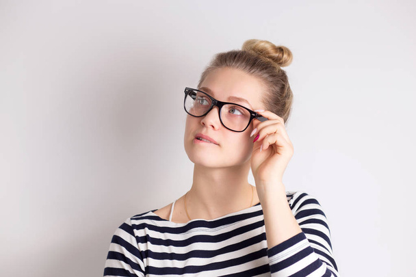 Indoor shot of cute woman in glasses looking away, having doubtful and indecisive face expression, pursuing her lips. Confused young female posing isolated at white wall with copy space. - Foto, Imagem