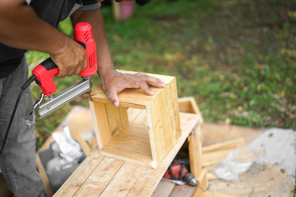Азиатский плотник стреляет гвоздь Air Nailer к деревянной квадратной коробке в саду на заднем дворе дома
 - Фото, изображение