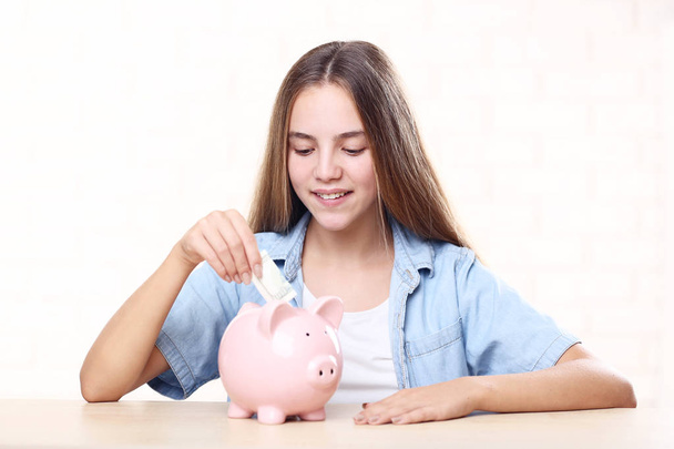Fiatal lány rózsaszín piggybank és a dollár bankjegyek asztalnál ül - Fotó, kép