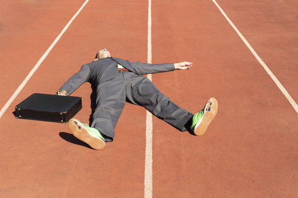 Un homme d'affaires épuisé en costume gris avec chemise et cravate vertes, mallette noire et chaussures de course vertes cassées allongées sur une piste de course
. - Photo, image