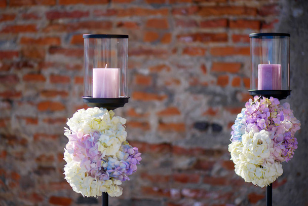 Minimalist outdoors two candle holders with pastel colored hydrangeas bouquets at the basis, decorative element for a formal event or a wedding, with a terracotta color brick wall in the background - Photo, Image