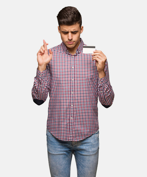 Young man holding credit card crossing fingers for having luck - Photo, Image