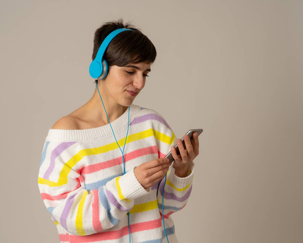 Portrait of gorgeous and happy brunette woman in colorful striped jumper listening music in headphones singing and dancing on neutral background. In positive emotions, youth and happiness concept. - 写真・画像