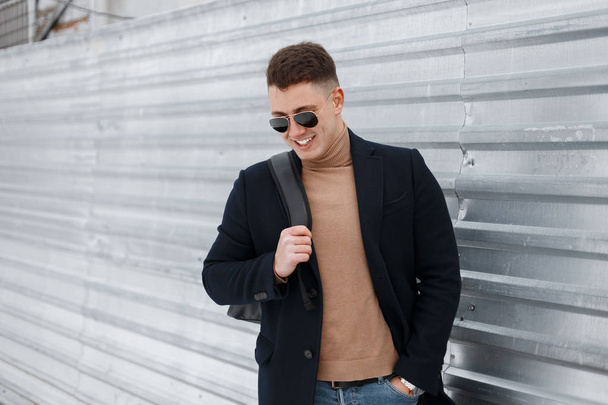 Stylish young hipster man in sunglasses in a stylish coat in jeans in a trendy knitted sweater with a backpack is standing and smiling near the metal fence. Cheerful guy travels. - Zdjęcie, obraz