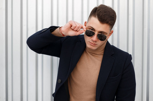 Portrait of a young attractive man hipster in stylish jacket in a vintage sweater with sunglasses with a stylish hairstyle near a metal wall. Handsome guy businessman. - Photo, Image