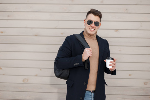 Ein fröhlicher junger Hipster mit stylischer Sonnenbrille in modischer Frühlingskleidung und trendigem Rucksack steht neben der Holzwand und hält draußen eine Tasse mit heißem Kaffee in der Hand. Lustiger Typ ruht sich aus. - Foto, Bild