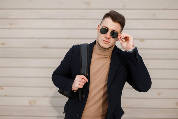 Привлекательный молодой хипстер в стильных очках в модном пиджаке в винтажном трикотажном свитере со стильной прической с рюкзаком на фоне деревянной стены. Американский парень
. - Фото, изображение