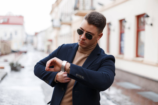 Jeune homme d'affaires élégant dans un manteau élégant en lunettes de soleil à la mode avec une coiffure élégante dans un pull tricoté regarde une main sur le fond des bâtiments vintage. Homme attrayant avec succès
. - Photo, image