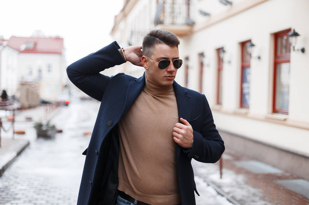 Attractive successful young hipster man in black stylish coat in black sunglasses in vintage knitted sweater posing in the city on the background of modern buildings. Handsome fashionable guy. - Foto, Imagem