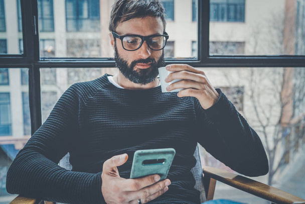 Bearded hispanic man in black pullover using mobile phone application for mobile blog. Confident young hipster guy read news via cellphone from social network at modern loft.Blurred background. - Φωτογραφία, εικόνα